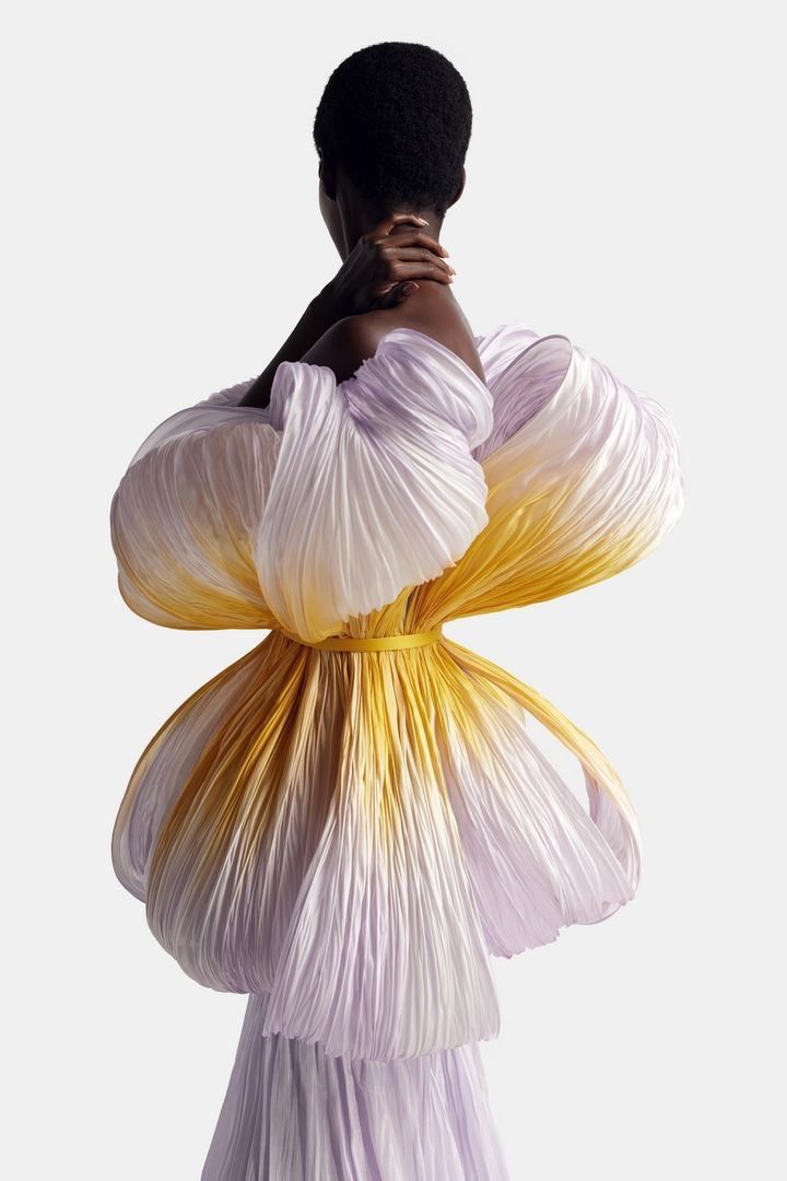 a black woman poses in a beautiful lavender and yellow ombré dress. the dress is pleated and  appears to be made from silk. it looks like it's made to look like flower petals.