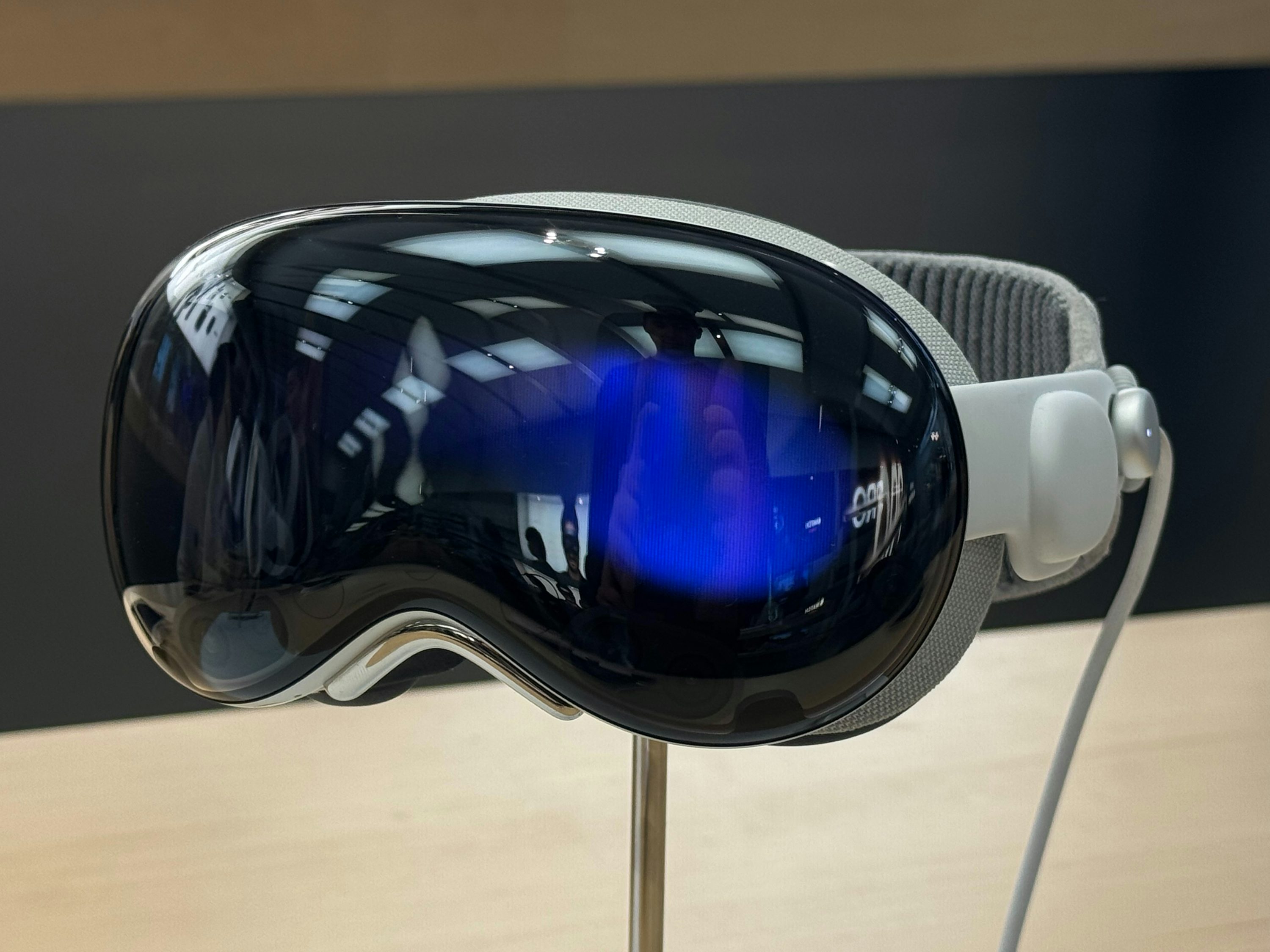 a closeup shot of a vr headset displayed on a metallic stand. the headset has a glossy black front panel with blue reflections visible. the strap is gray and textured, and the sides are white. a white cable is attached to the side. the headset is sitting on a lightcolored surface with a dark background behind it.
