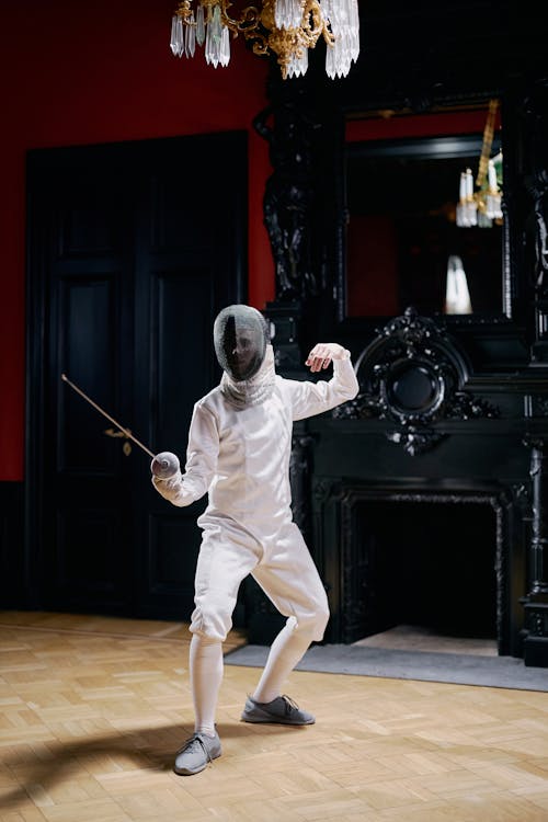 a fencer, dressed in a white uniform and mask, stands in a poised stance inside an elegant room with a wooden floor.  the room features a large dark fireplace and an ornate chandelier. the fencer holds a foil, ready for action.  the scene is captured in a still image that conveys both elegance and athleticism.