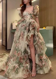 a beautiful woman is wearing a floral patterned dress and high heels. she is standing indoors and posing for the camera. the dress is a high low style with ruffles, and she is wearing a delicate necklace.