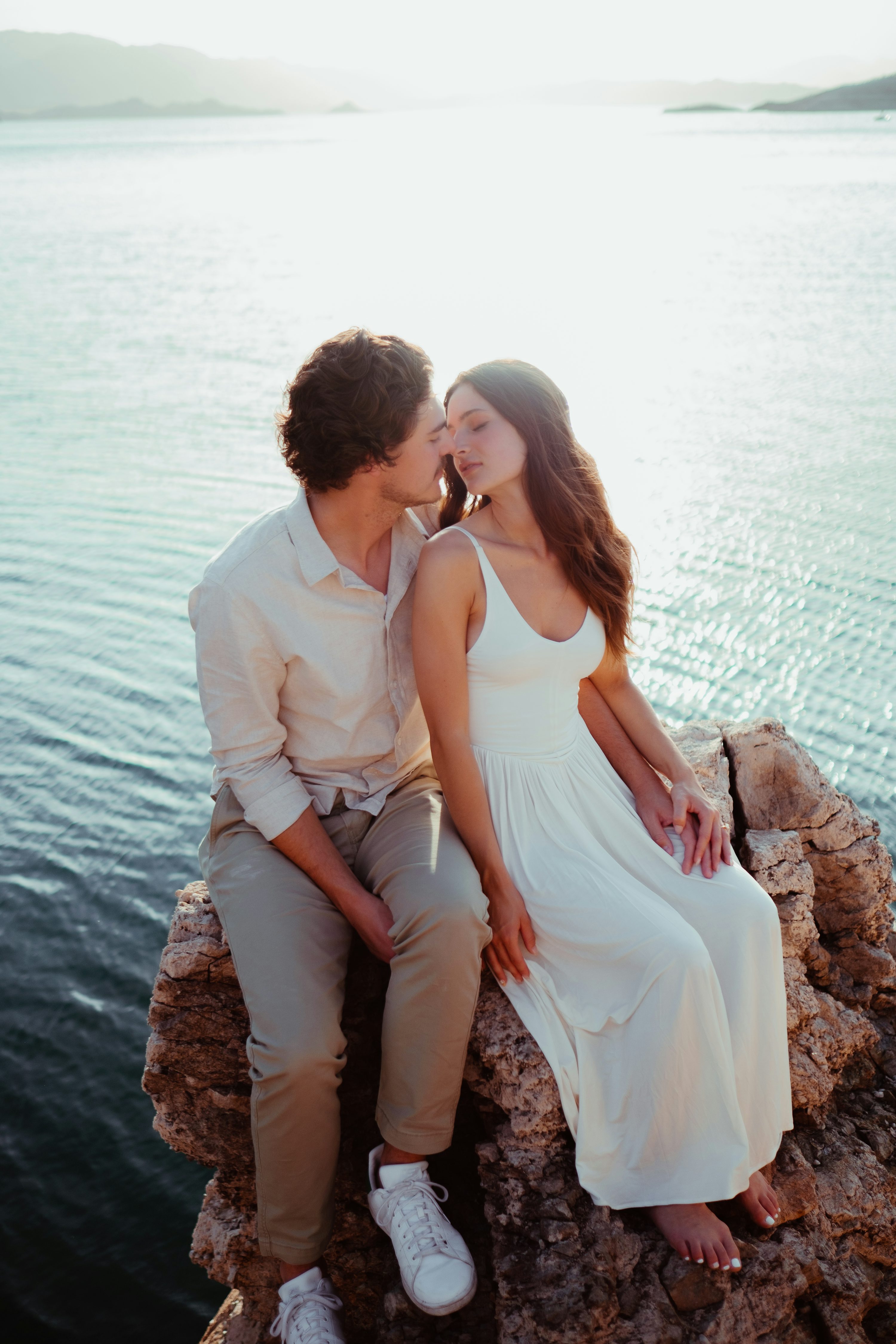 a young couple is sitting on a rocky outcrop by the ocean, embracing and sharing a tender kiss. the woman is wearing a flowing white maxi dress, and the man is wearing beige pants and a lightcolored shirt. they are basking in the warm glow of the setting sun. the scene is peaceful and romantic, showcasing the beauty of the natural landscape and the intimacy of their relationship.