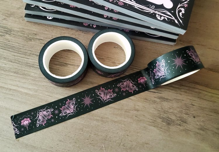 a closeup shot of two rolls of black washi tape with a moth and floral design, next to a partially unrolled strip of the same tape and a stack of black notebooks, all resting on a wooden surface. the tape features pink moths, stars, and flowers.