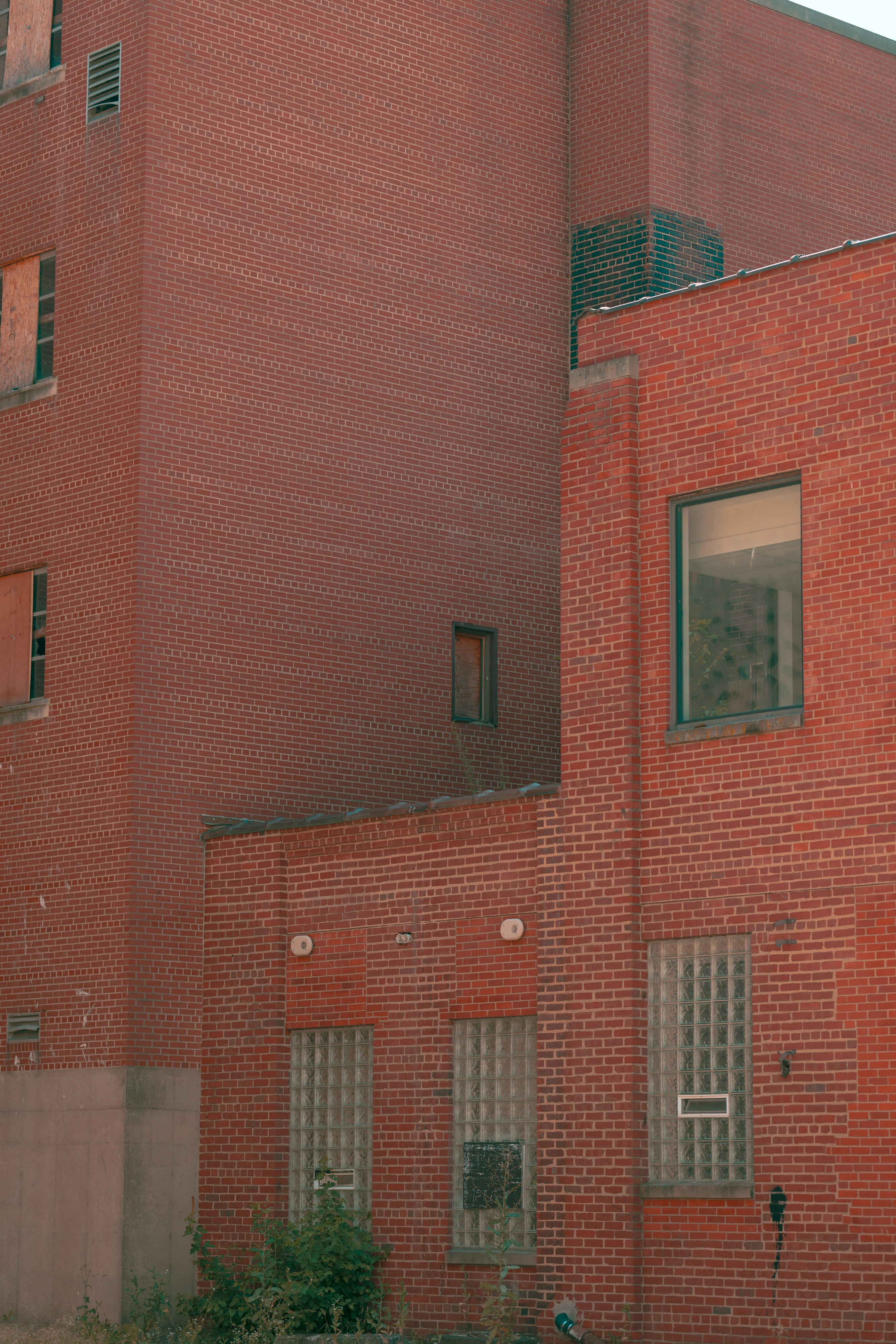 the image features a segment of a large, multistory brick building. the building, constructed from red brick, features multiple windows of varying sizes and styles. some windows are boarded up with metal grating, hinting at possible disuse or security measures.  the overall aesthetic leans towards an industrial or urban setting. the natural daylight casts subtle shadows on the building's facade, adding depth and texture to the scene.  a glimpse of greenery at the base suggests a touch of nature amidst the urban environment.