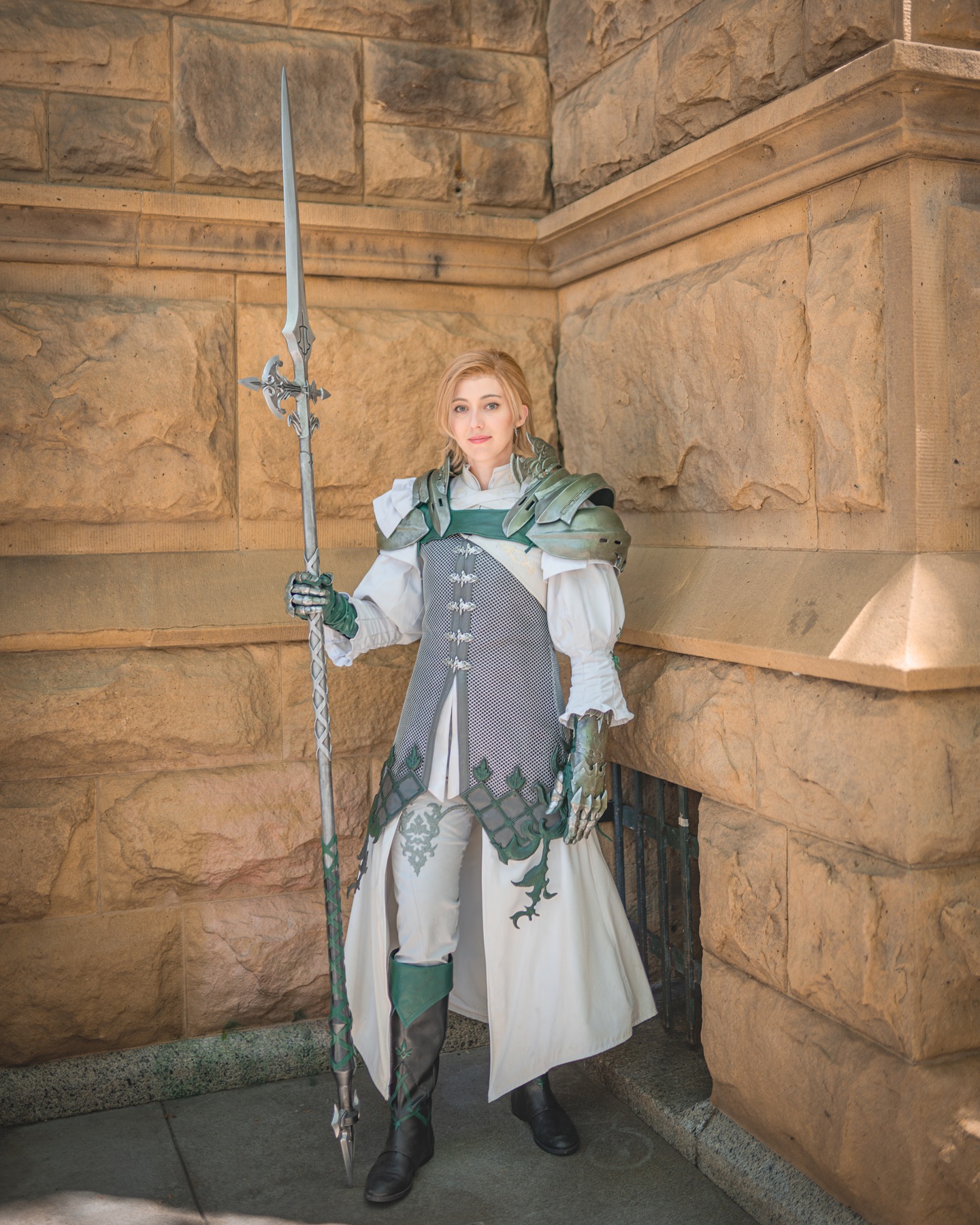 a person in cosplay, dressed as a female warrior from a fantasy game, possibly final fantasy xiv. the cosplayer is wearing an elaborate costume with armor, a long white coat, and green accents. they are holding a long spear and standing against a stone wall. the setting appears to be outdoors, and the image is a fullbody portrait.