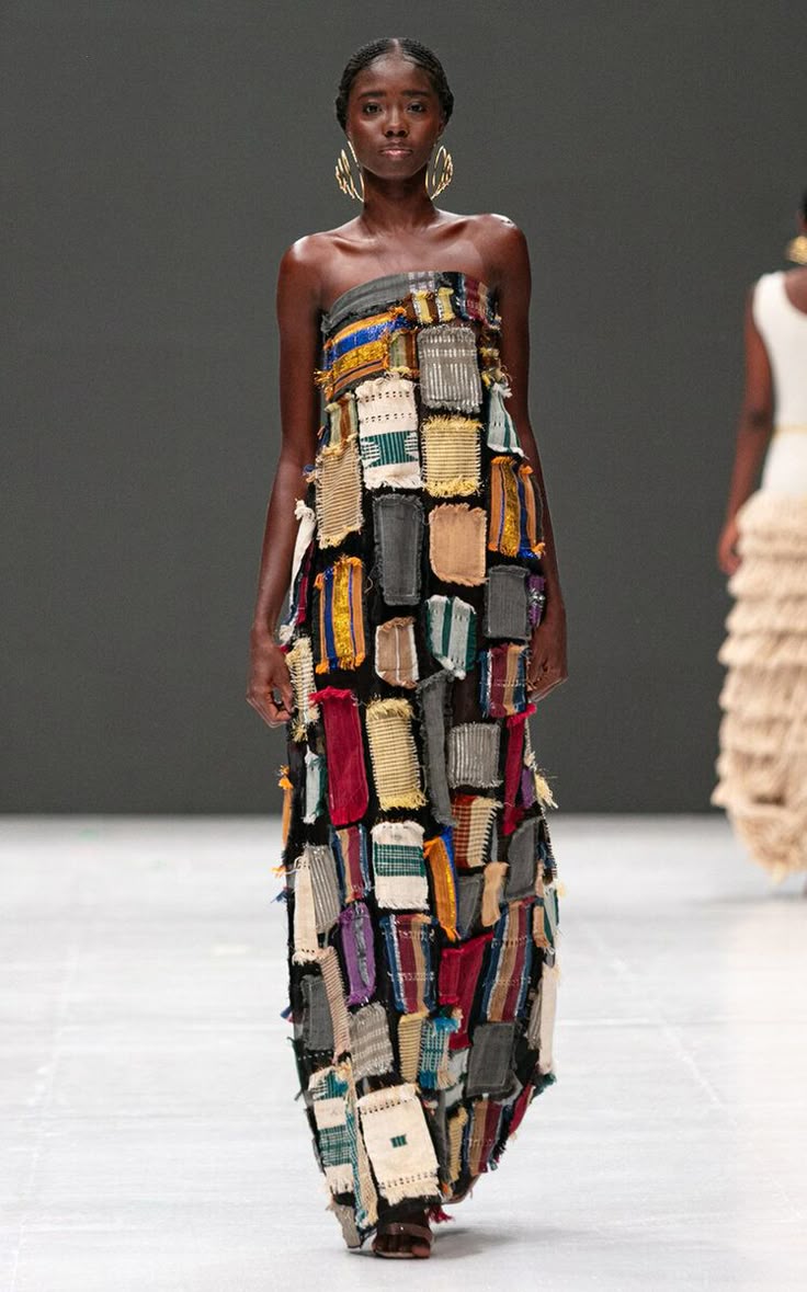 a black female model is walking on a runway in a strapless, floorlength dress made of rectangular patches of different colors and textures. she is wearing large, gold earrings. another model, partially visible, is behind her.