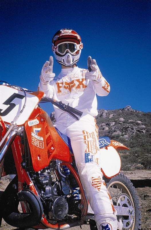 a motocross rider in full gear is standing beside a dirt bike with his hands in the air. the rider is wearing a white fox racing jersey with the company logo, white pants, and a red and white helmet. he is also wearing goggles, gloves, and boots. the dirt bike is a honda cr250 in red and white. the rider is posed against a background of rocky hills and a bright blue sky.