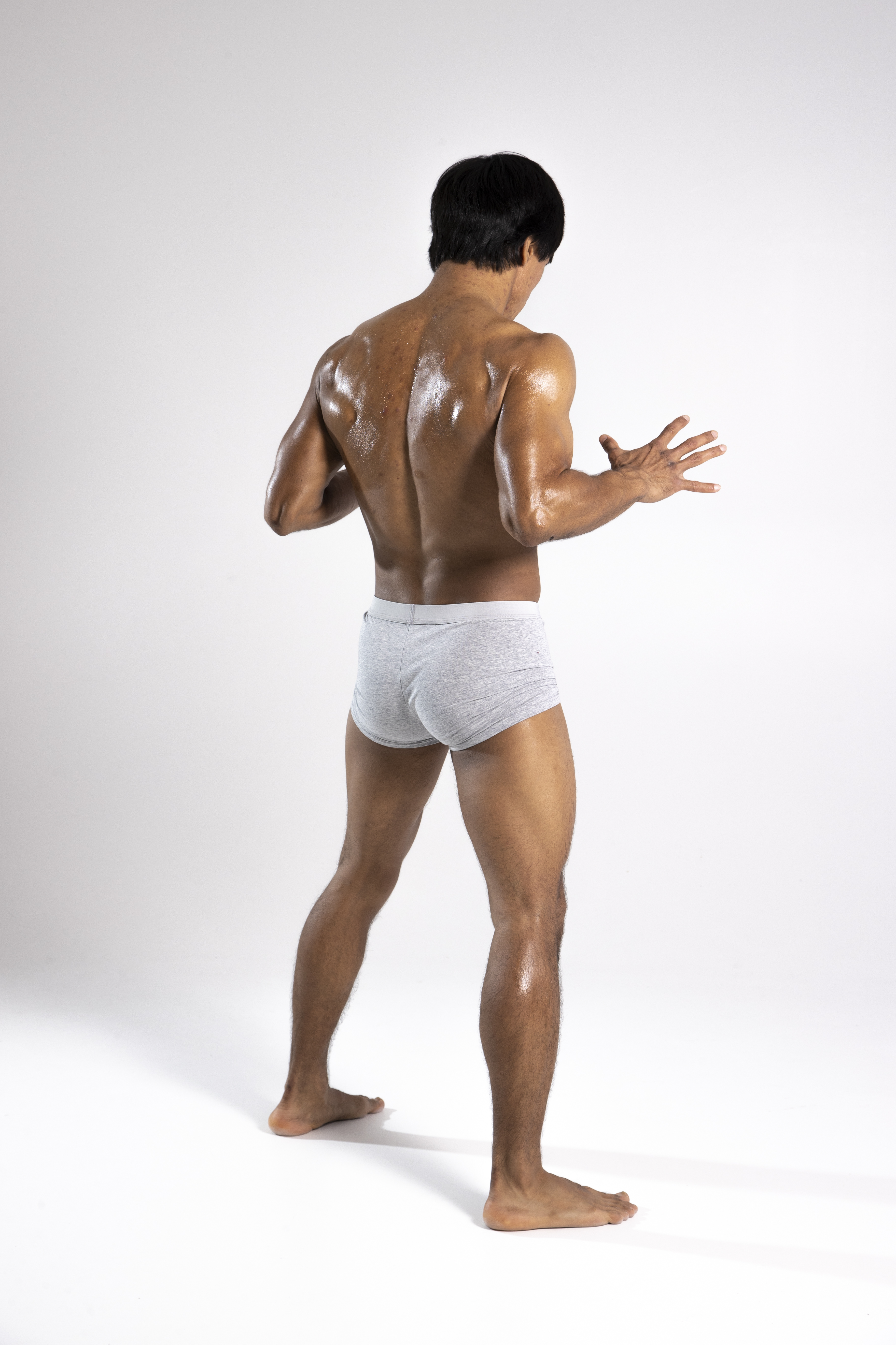 a man stands with his back to the viewer in a studio setting. he has dark, short hair and is wearing gray underwear. his torso and legs appear wet and are glistening, possibly from sweat or oil. his arms are slightly raised and bent at the elbows with his hands open, palms facing forward. the background is a plain white backdrop, which highlights the man's body and posture.