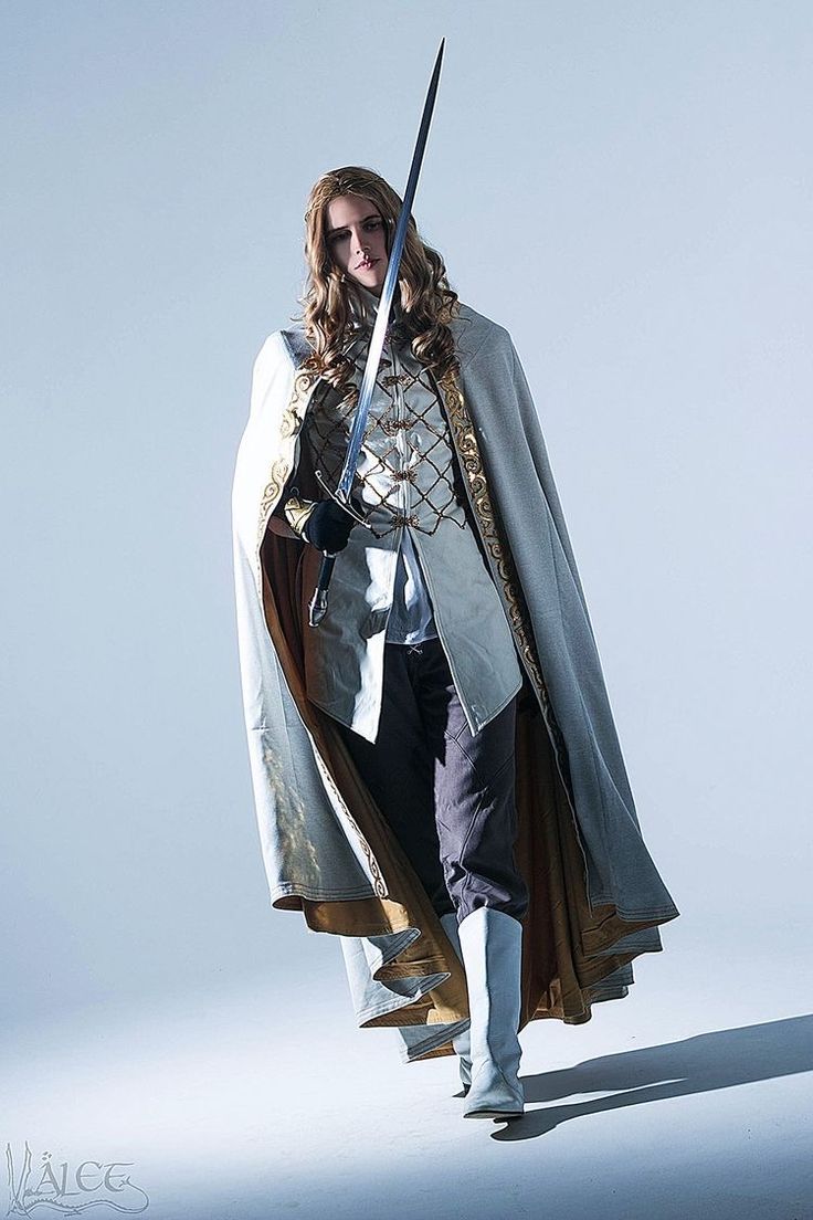 the image is a professional studio shot of a male cosplayer, possibly inspired by lord of the rings or similar high fantasy settings. he is elegantly dressed in a flowing lightblue/white cloak with gold detailing, a matching tunic and pants, and lightblue boots. he carries a silver sword across his shoulder. his long blonde hair is loose. the overall aesthetic is ethereal and aristocratic. the background is a plain lightblue backdrop, drawing focus to the cosplayer's costume and pose. the lighting is dramatic, with shadows adding depth to the image.  the watermark “välee” is visible in the bottomleft corner.