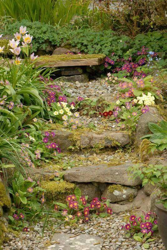 a charming garden path leads up a gentle slope, featuring natural stone steps covered in moss and surrounded by vibrant clusters of flowers, predominantly primroses in shades of pink, red, and yellow. the path is composed of small gravel, and a rustic wooden bench sits nestled amongst the greenery at the top. the scene is lush and verdant, with various plants and foliage adding to the overall natural beauty of the garden.