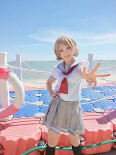 a young woman is cosplaying at the beach. she is wearing a japanese school uniforminspired outfit with a sailor top, a pleated skirt, and kneehigh socks. she is posing with a peace sign and smiling brightly. the background features a clear blue sky, sparkling ocean water, and a red and blue floating platform.