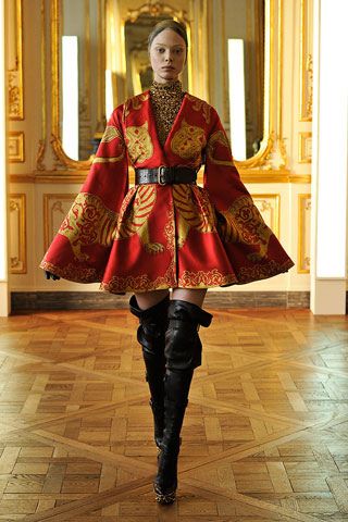 a full shot of a fashion model wearing a striking red and gold dress adorned with a tiger print.  the dress features wide sleeves and a belted waist.  she's wearing tall black boots and is posing indoors on a polished wooden parquet floor in an ornate setting, possibly a palace or grand mansion.  the overall aesthetic is one of high fashion and luxury.