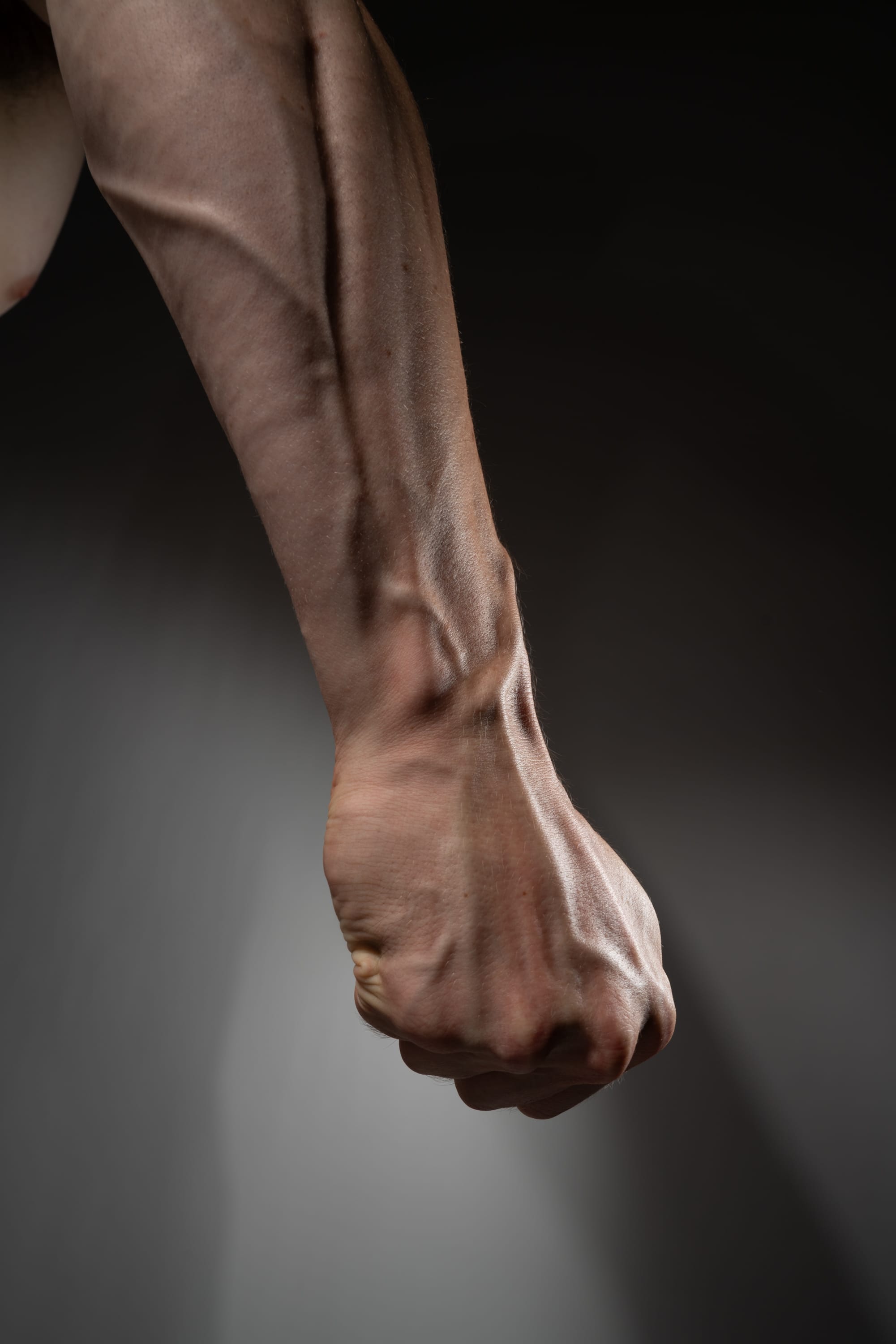 a closeup side view of a muscular man's arm and clenched fist, set against a dark gray background. the image highlights the definition and vascularity of the arm, emphasizing strength and physical power.