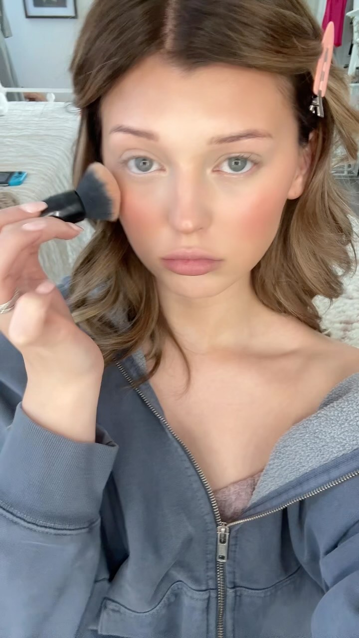 a woman with blonde hair and light eyes applies makeup to her face with a brush. she is wearing a gray hoodie with a zipper and has hair clips in her hair. she is also wearing a ring on her finger.
