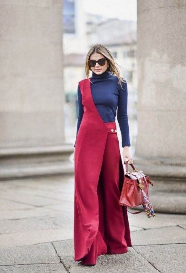 a woman stands on a stone street in front of a stone building. she is wearing a blue turtleneck sweater with a red jumpsuit on top, sunglasses, and carrying a red bag.