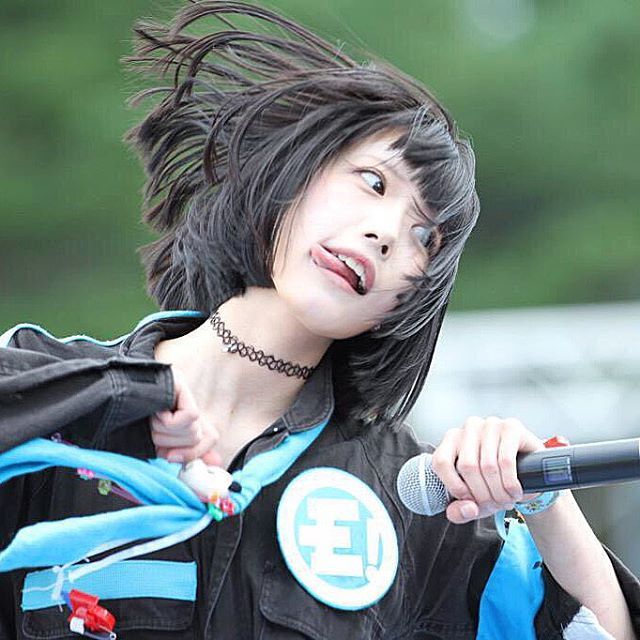 a young asian woman with black hair and a blue scarf is singing into a microphone while looking up and smiling. she is wearing a black shirt and a black jacket with a white and blue patch that says "e!" on it. her hair is styled in a short, choppy cut and is being blown by the wind. she is wearing a black choker necklace and has a vibrant blue ribbon on her right wrist. she appears to be performing on a stage or at an outdoor event, and she seems to be enjoying herself. she is surrounded by a lively atmosphere.