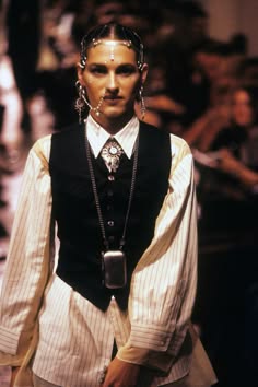 a fashion model wearing a white striped shirt with a black vest, accessorized with large silver earrings and a silver chain necklace with a silver pendant. the model is posing seriously, looking at the camera, with a blurred background of other models and audience members.