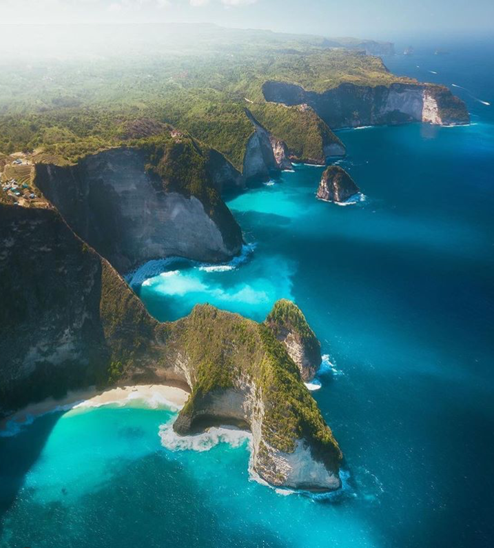 an aerial view of a tropical coastline featuring a series of rocky cliffs covered in green vegetation. the cliffs meet the turquoise water of the ocean, creating white waves where they collide. there is a small beach nestled between the cliffs. the scene is bright and sunny, highlighting the vibrant colors of the landscape. the location appears to be nusa penida, specifically kelingking beach.