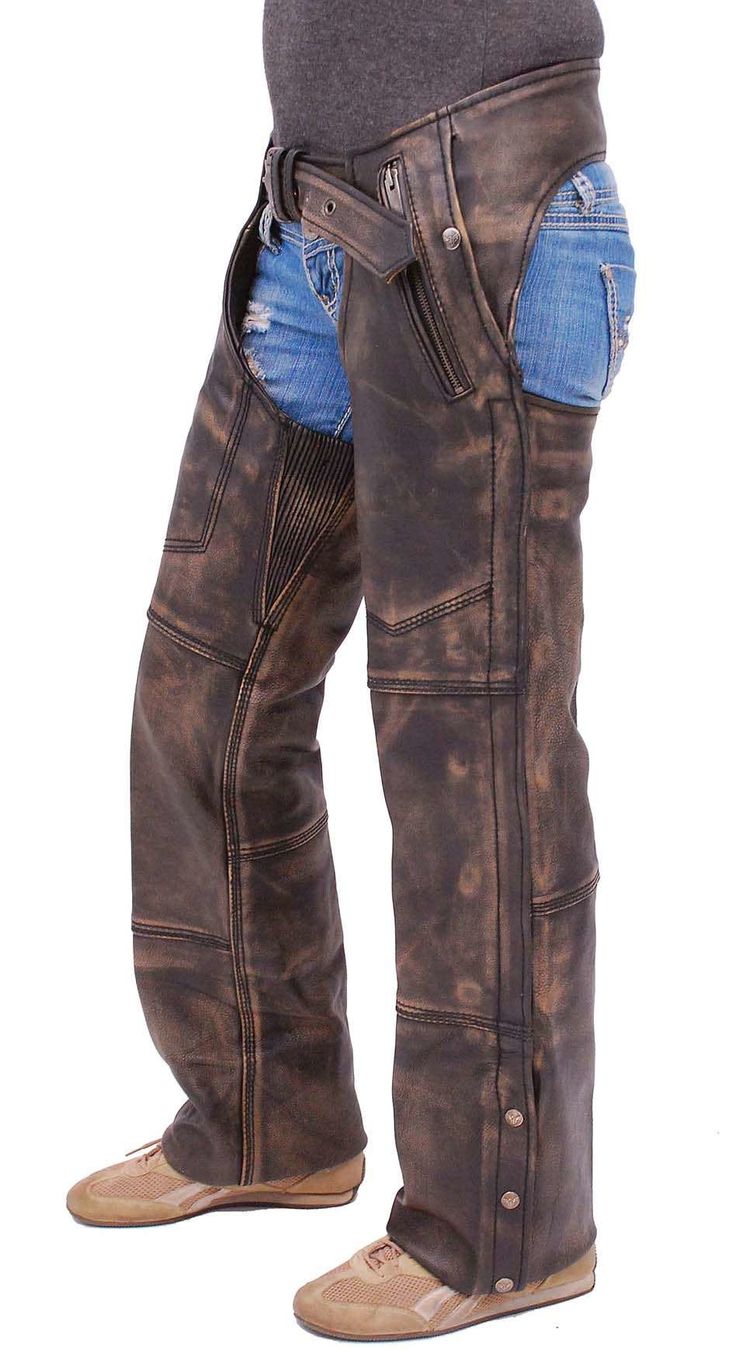 a full shot of a woman wearing brown distressed leather chaps over blue jeans. she is wearing tan sneakers. the chaps are detailed with stitching and have a belt.