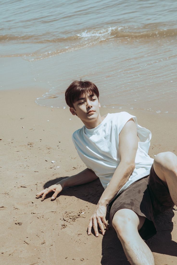 a young man with short brown hair and a white tshirt is posing on a beach. he is lying on his side in the sand, with his left arm outstretched and his right arm bent. the ocean can be seen in the background. the photo is taken from a high angle, and the lighting is natural and sunny.