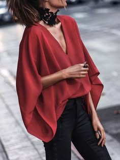 a woman wearing a red blouse with a black choker and black jeans. she is standing on a street and looking at the camera.