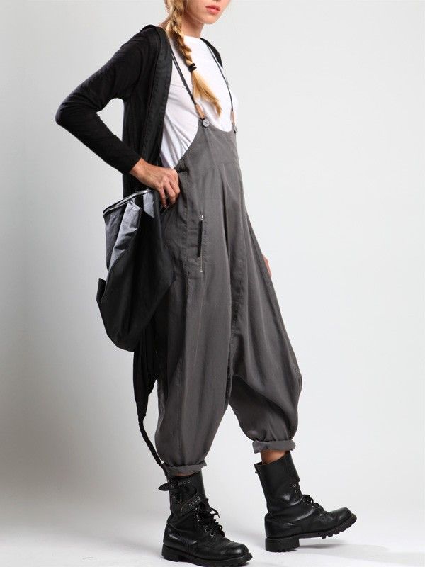 a woman is standing in front of a white background. she is wearing a pair of gray overalls, a white tshirt, and a black cardigan. she has a black backpack over her shoulder. the overalls are cuffed at the ankles and she is wearing black boots. her hair is braided.