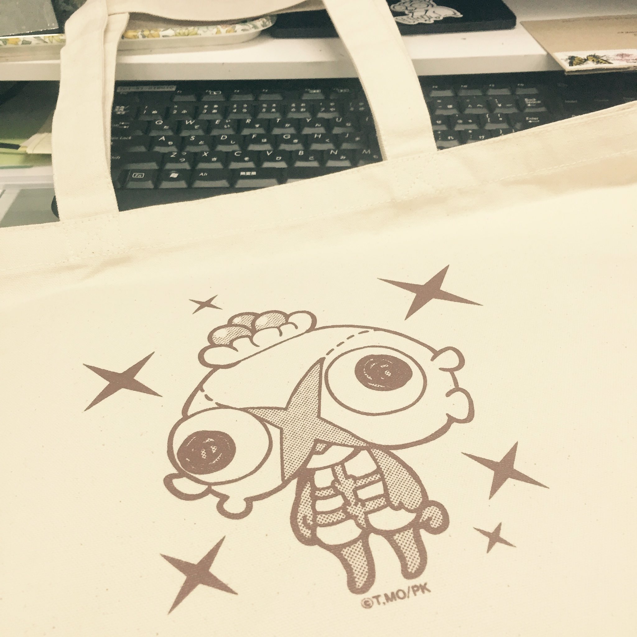 a creamcolored tote bag with a cute, brown cartoon character printed on it. the tote bag is sitting on a desk with a keyboard in the background.