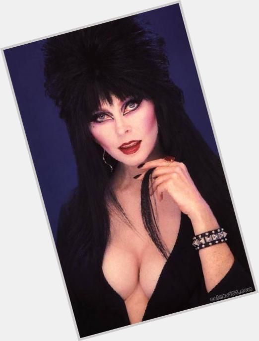 a close up portrait of a woman with long dark hair wearing a black dress with a low cleavage, showing off her jewelry and makeup.