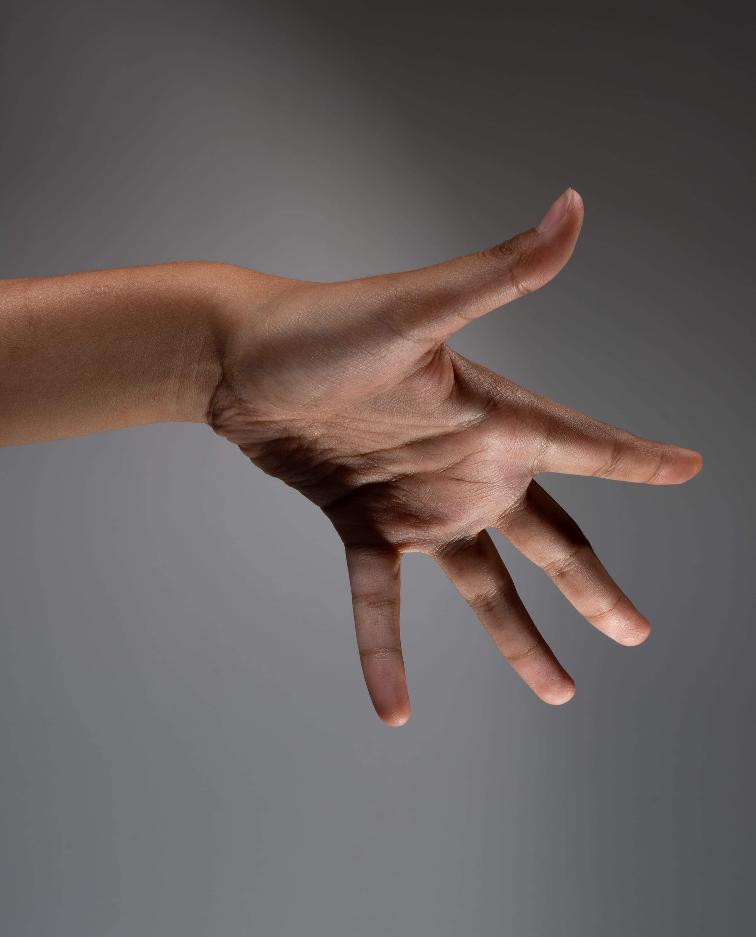 a hand is extended against a gray background. the hand is outstretched, with the fingers slightly spread apart. the light source is from above and to the left, casting shadows on the hand and arm. the image is taken from a side view, focusing on the hand and arm.