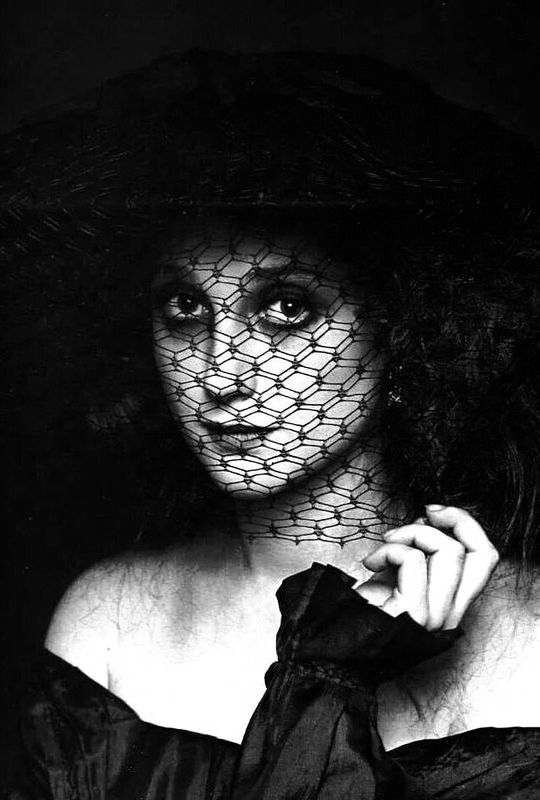 a black and white portrait of a woman wearing a black hat and veil. she is looking directly at the camera, her eyes are visible through the net. she is dressed in a dark outfit and has a dramatic and mysterious look. the lighting is soft and the background is dark.