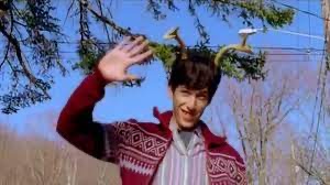 a man with antlers on his head is smiling and waving at the camera. he is wearing a red sweater and is standing in a forest. the trees are bare and there is snow on the ground. the man is looking at the camera and seems happy.