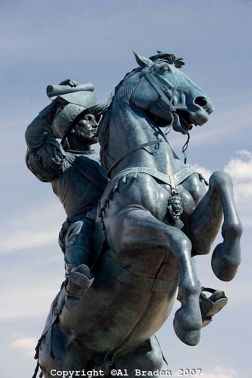 a closeup view of a bronze equestrian statue, possibly depicting a spanish conquistador.  the statue is weathered and shows fine details in the sculpting of both the horse and the rider. the rider is shown holding a rolledup document, and wears what appears to be a helmet and ornate clothing. the horse is captured midstride, with its legs raised. the background is a partly cloudy sky. the image has a copyright notice at the bottom.