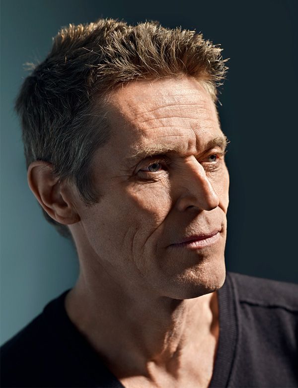 a portrait of actor willem dafoe. he is a mature man with short grey hair, wrinkles, and is wearing a black vneck shirt. he is looking away with a slight smile. the photo is a closeup headshot taken in a studio setting.