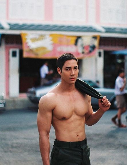 a young, shirtless, muscular man stands on a city street. he is looking off to the side with a slightly mischievous grin.  he is wearing a dark green military style cargo shorts. his black hair is slightly tousled and he has a dark jacket draped over his shoulders and in his hand. there are shops in the background.