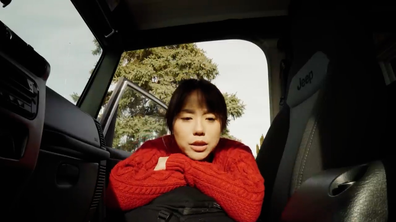 a young woman with shoulder length black hair is sitting in the driver's seat of a black jeep with the door open. she is wearing a red cable knit sweater and has her arms resting on the center console of the car. she has a neutral expression on her face. the interior of the car is black and the exterior is visible through the open door. the background features green trees and the lighting suggests that it is a bright and sunny day. the image is captured from a low angle.