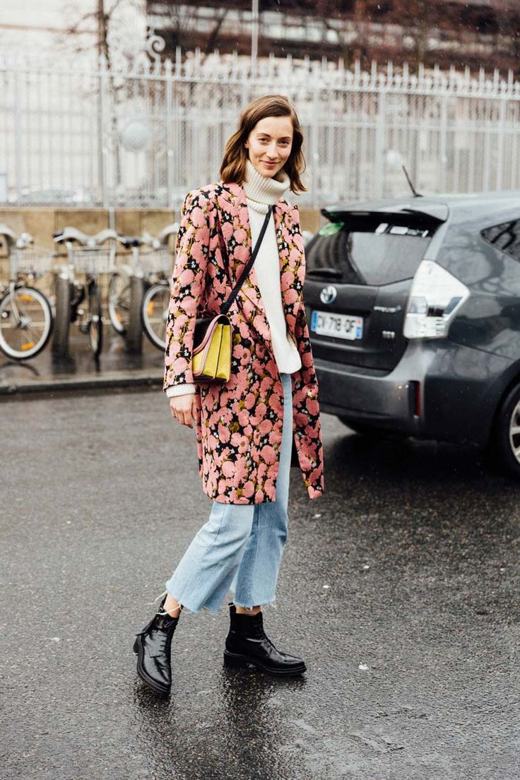 a stylish woman walks down a parisian street, dressed in a pink floral coat, light blue cropped jeans, black ankle boots, and a yellow and black handbag. she is wearing a white turtleneck sweater underneath her coat.  the setting is slightly rainy, with bicycles visible in the background, and a grey car parked nearby. the overall aesthetic is chic and fashionable, representative of parisian street style.
