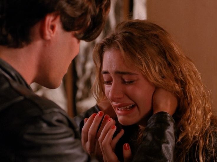 a woman with long, curly hair is crying in the arms of a man. she has tears in her eyes and her mouth is slightly open. the man is comforting her and holding her close. the image is closeup, with the focus on the woman's face. they are both wearing black leather jackets. the background is blurry, suggesting that the image is from a film or television show. the scene depicts a moment of emotional intimacy and sadness.
