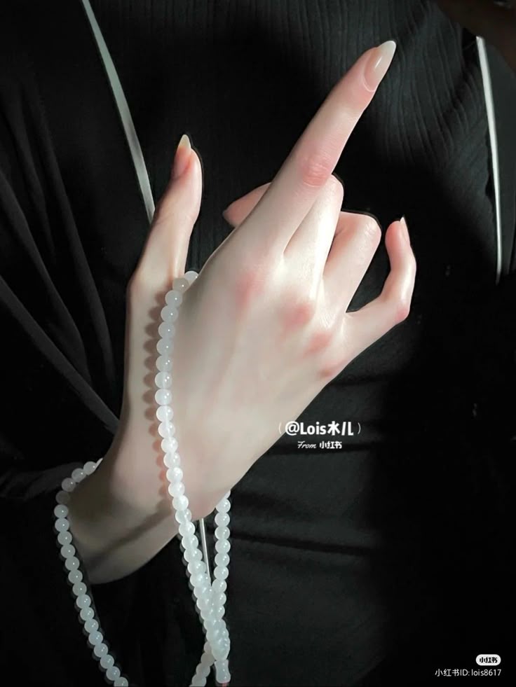 a closeup shot shows a pair of hands holding a strand of white beaded jewelry, set against a backdrop of black clothing. the fingers are elegantly extended, displaying long nails with a natural look. the hands are fairskinned, with subtle pink hues on the knuckles and fingertips. the jewelry consists of small, round white beads that cascade down from the hand. the black clothing provides a stark contrast to the fair skin and white beads, drawing focus to the hands and jewelry.