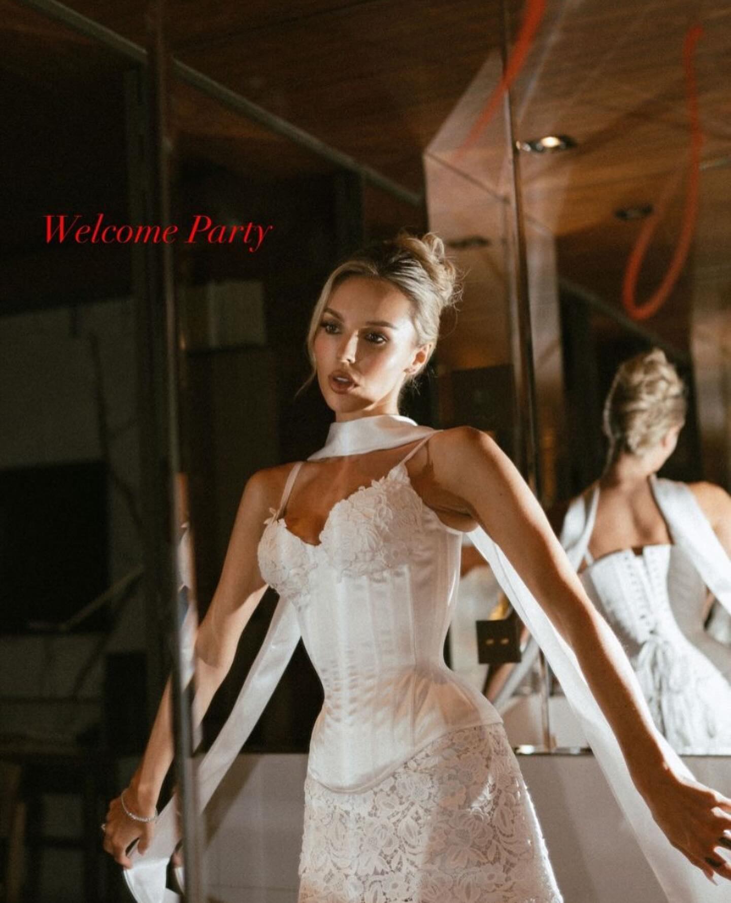 a beautiful woman with long blonde hair in a white lace corset and white skirt is looking directly at the camera. she is standing in a luxurious room with red light reflections. she is smiling and her eyes are sparkling.