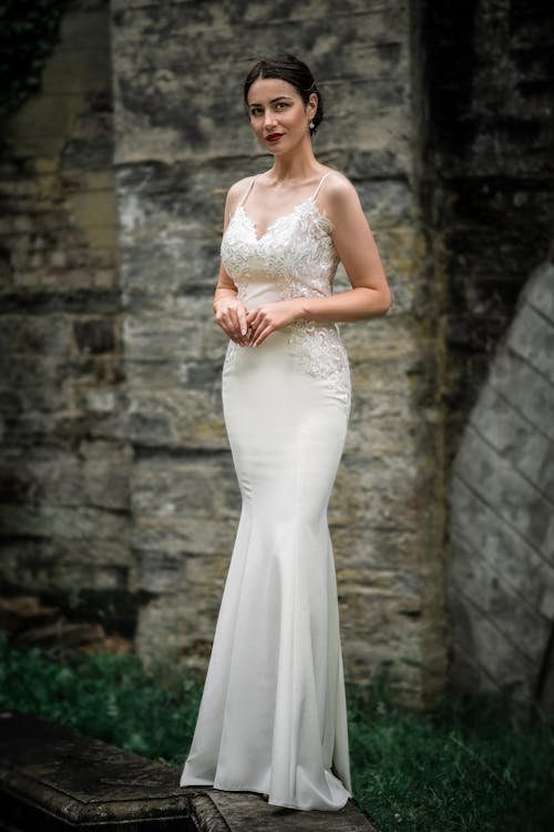 full shot of a beautiful bride in a white mermaid wedding dress. she is standing outdoors against a stone wall, her hands clasped together.  the dress has lace detailing on the bodice. she is looking directly at the camera with a confident expression. the background is blurred, highlighting the bride and dress. the overall mood is elegant and sophisticated.