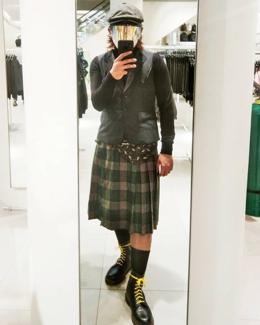 a man poses for a mirror selfie in a clothing store changing room. he is stylishly dressed in a gray vest, black turtleneck, green and black plaid kilt, black socks, and black laceup boots with yellow stitching. he is also wearing a black newsboy cap and a futuristic silver face shield. the environment suggests he is trying on the outfit, possibly considering a purchase.