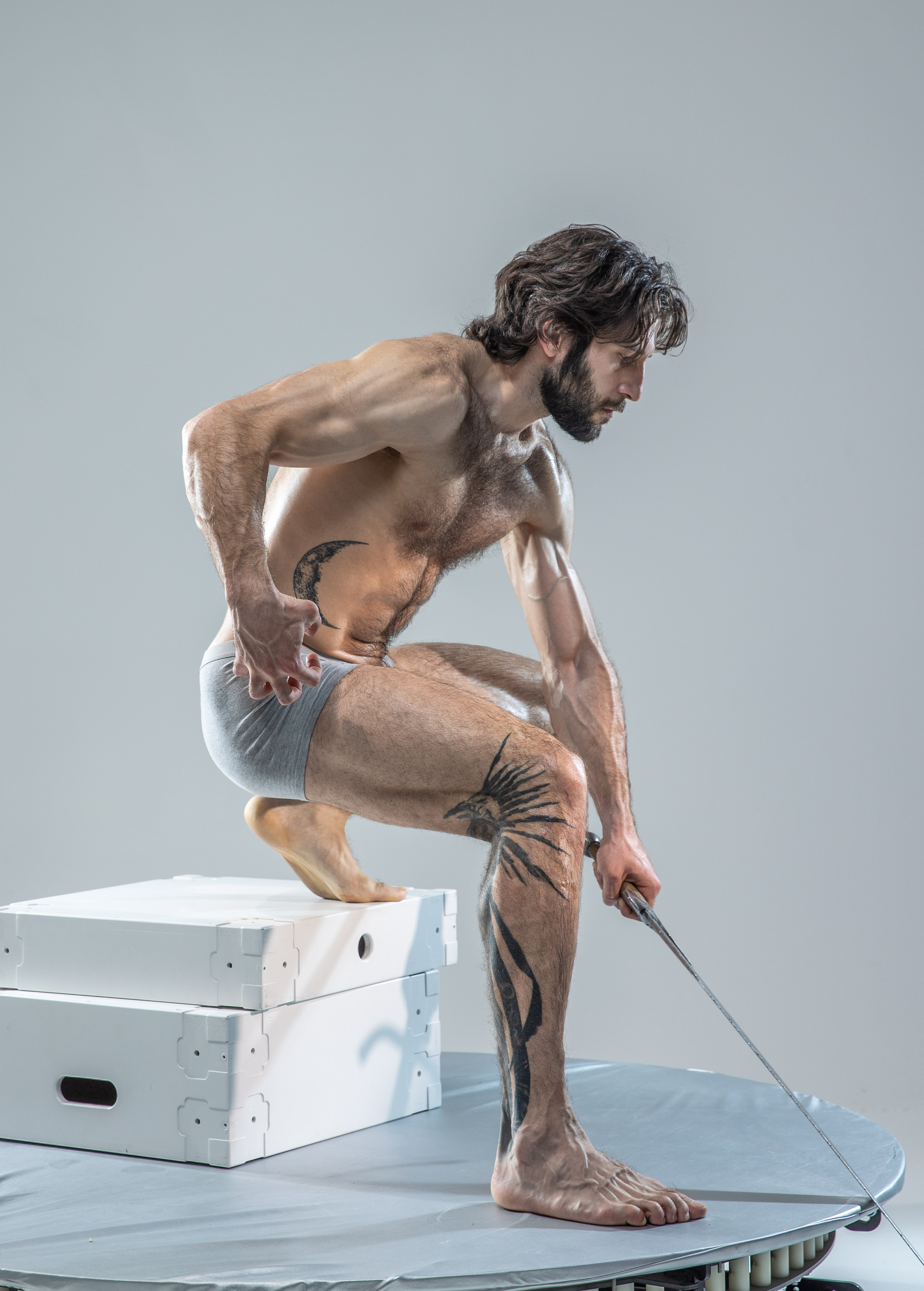 a muscular, athletic man with long hair and tattoos poses on a white platform in a studio setting. he holds a sword in his right hand, extending it outwards. he's shirtless, wearing only gray underwear, and is positioned with one foot on a white box and the other on the platform. the lighting is soft and diffused, characteristic of a studio environment.