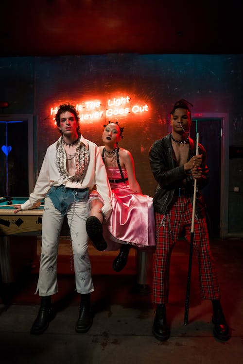 three stylish individuals pose in front of a pool table in a dimly lit room with a neon sign that says "hope is the light that never goes out". the person in the center is wearing a pink dress, while the other two are wearing more androgynous outfits. the room has a grungy, alternative vibe, and the image has a candid feel.  the overall aesthetic evokes a sense of rebellion and coolness.