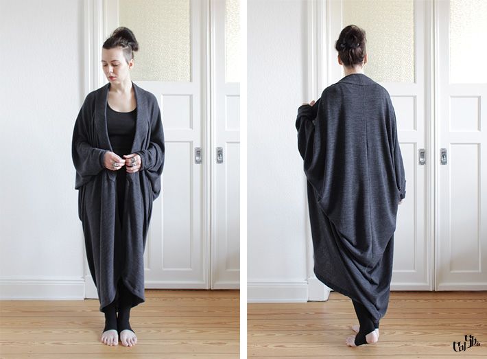 two images side by side show a woman modeling a gray oversized cardigan and leggings. the left image shows her from the front, looking down, with her hands slightly raised, wearing a gray cardigan over a dark top, paired with leggings and barefoot. the right image shows the same woman from the back, highlighting the cocoon silhouette of the cardigan, which is long and voluminous. both images are taken indoors against a backdrop of white doors and a wooden floor. the woman has her hair styled in a bun.