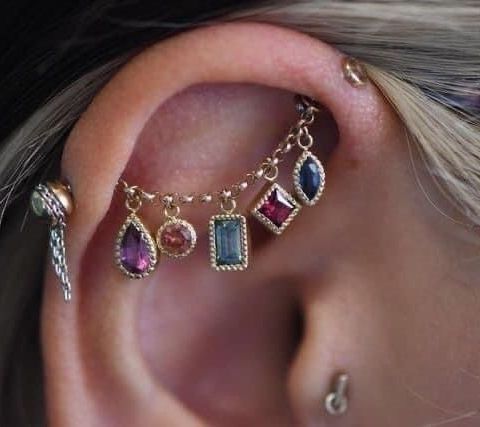 loseup view of a person's ear with multiple piercings. the main focus is on a helix piercing adorned with a delicate gold chain featuring various small gemstones of different shapes and colors (ruby, sapphire, tourmaline). the gemstones are set in gold settings, each hanging from the chain.  there's also a small stud piercing visible lower on the earlobe. the overall style is delicate and bohemian.  the background is slightly blurred, drawing attention to the piercing and jewelry.