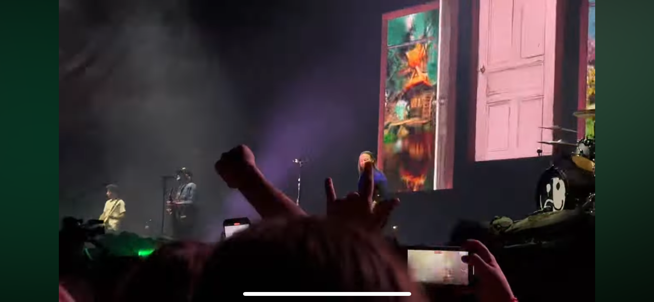 a shot from the audience at a concert. there is a band on stage, playing music for a crowd of fans. the people in the audience are enjoying the show, some of them are holding their phones up to record the event.  the band is performing in front of a large screen with a door as the background.