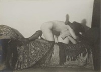the black and white photograph depicts a nude couple engaged in an intimate act on a bed. the photograph is somewhat blurred and suggestive, rather than explicitly graphic.  the couple's bodies are intertwined, with the top figure appearing to be a man and the lower figure a woman.  the woman's legs are clad in stockings. they are lying on a bed covered with a richly patterned paisley or similar design fabric, with a dark fringed shawl or blanket partially draped over them.  the overall mood is sensual and mysterious, with a focus on the emotional intimacy rather than precise anatomical detail. the style hints at early to mid 20thcentury art photography. the slightly blurry nature of the image could be intentional or a result of the vintage photographic techniques.