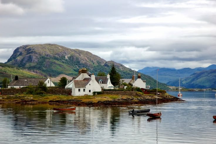 the image showcases a picturesque coastal village nestled amongst rolling green hills and mountains in the scottish highlands.  whitewashed houses cluster together on a small peninsula jutting into a calm loch.  several small boats and a sailboat are moored in the tranquil water. the sky is overcast, adding to the serene and atmospheric feel of the scene. the overall impression is one of peacefulness and natural beauty.