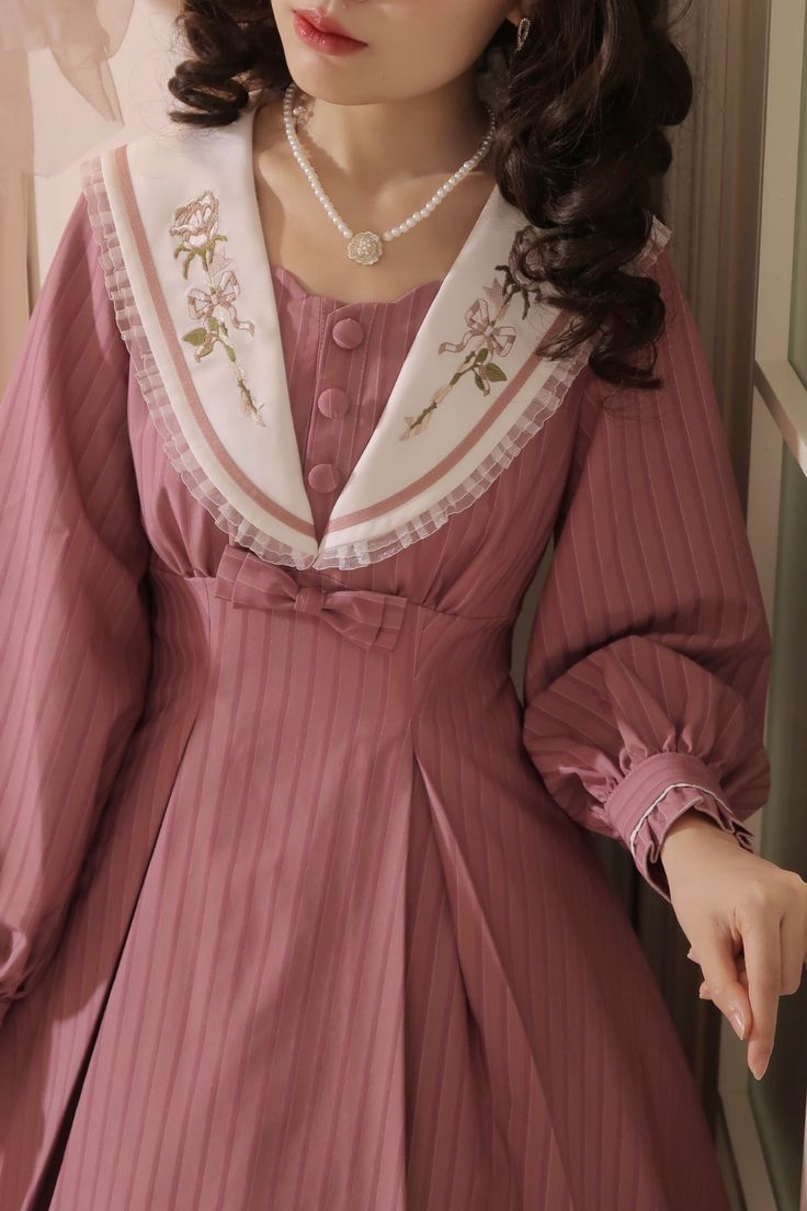 a closeup shot of a woman wearing a pink lolita fashion dress. the dress features a square neckline, a sailor collar with floral embroidery, long sleeves with ruffled cuffs, and a pleated skirt. the dress is cinched at the waist with a matching fabric belt. the woman is also wearing a pearl necklace and has her hair styled in loose curls.