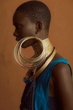 a portrait of a woman with a striking golden necklace and earrings. she's looking to the side and the background is a warm, brown. her skin is dark and beautiful, she has a short haircut and a look of contemplation.  the image is artful and stylish.