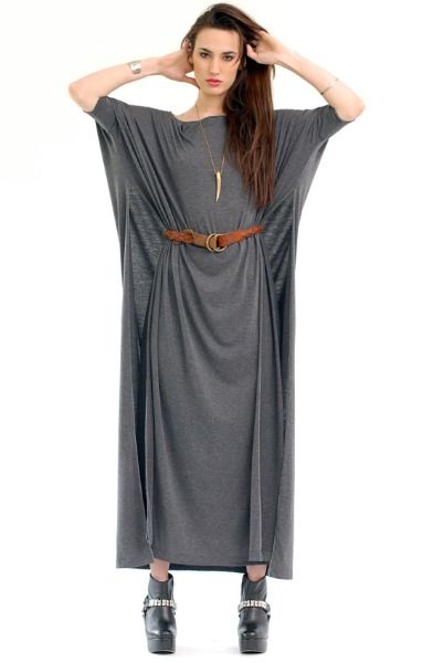 a woman stands facing forward, wearing a long, gray, oversized dress cinched at the waist with a brown belt. she has long, dark hair and is wearing a long necklace with a pendant. her arms are raised, with her hands near her head. she is wearing black platform boots. the background is white.